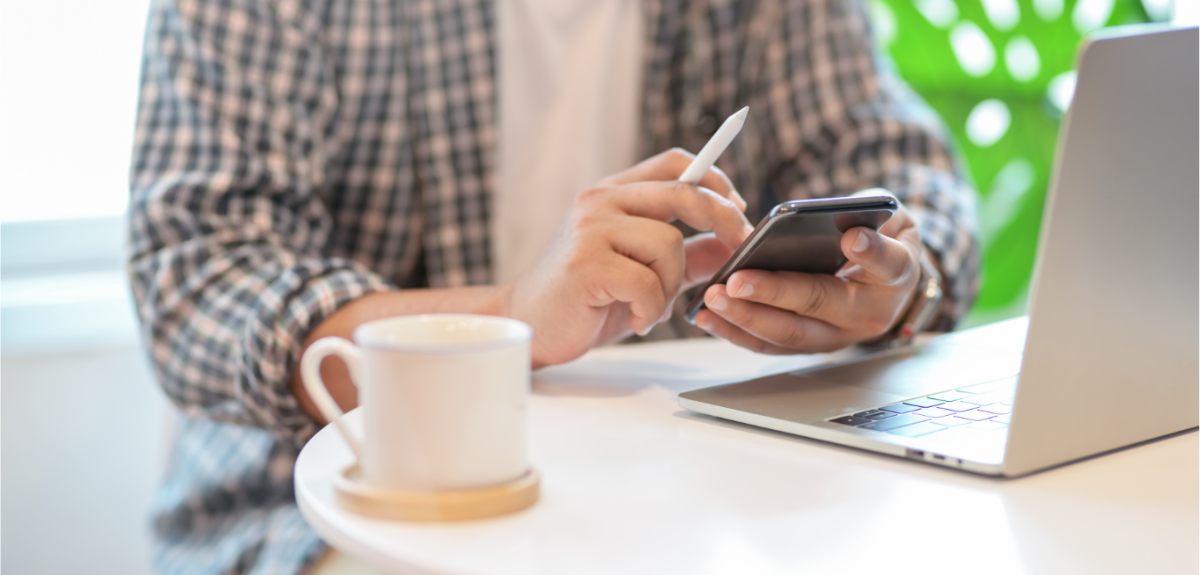 A person using a phone and laptop at a table with a cup of coffee